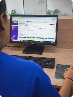 A person in a blue shirt is using a desktop computer at a desk, viewing a web application with data entry fields on the screen. The keyboard and mouse are on the desk.
